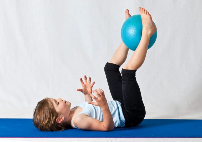Das Kind liegt auf einer Isomatte und streckt die Beine in die Höhe. Mit den Beinen hält es einen Ball in der Luft.