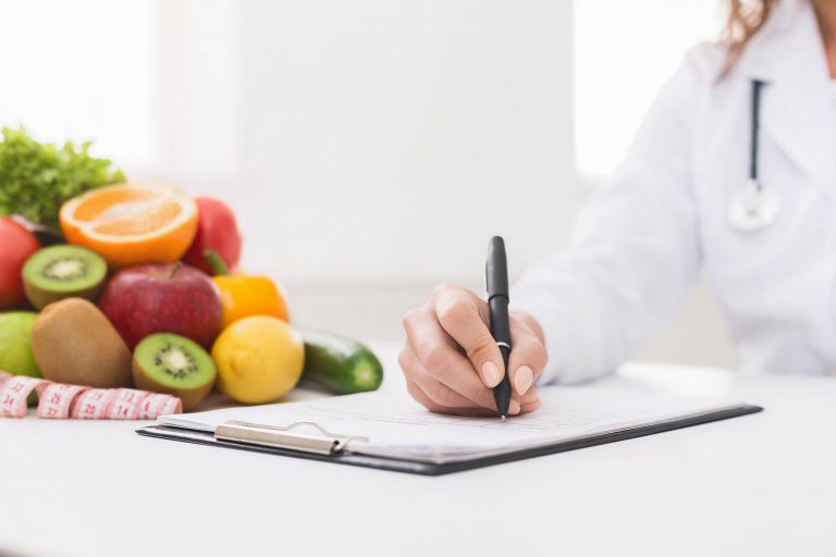Nahaufnahme einer Frauenhand, die mit einem Kugelschreiber auf Papier schreibt. Links sind Obst und Gemüse zu sehen.